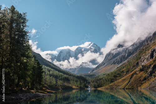 Scenic landscape with turquoise mountain lake in autumn valley against large snow mountains in low clouds in morning sunlight. Alpine lake with view to sunlit high snowy mountain range in low clouds.