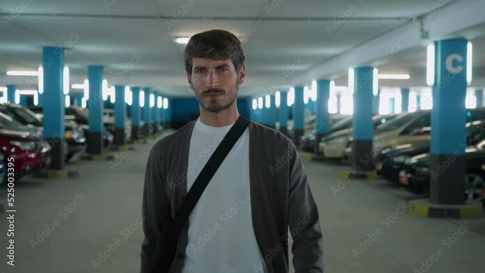 Young guy walks through the underground parking level and looks at the camera, looking for a rental car.
