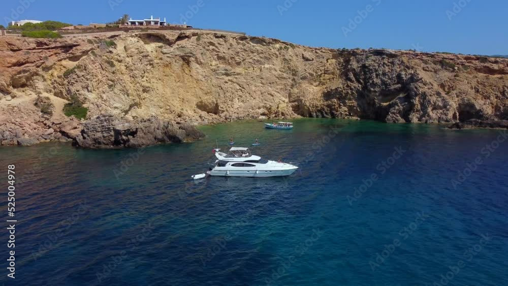 White luxury yacht in bay on cliff villaButtery soft aerial view flight fly backwards drone footageof cliff edge beach Sa Figuera borda Ibiza summer 2022
