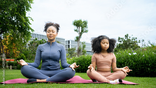 African american Mother daughter doing yoga exercises on grass in park at day time. People having fun outdoors. Concept of friendly family and of summer vacation. Mom and child in the lotus position.