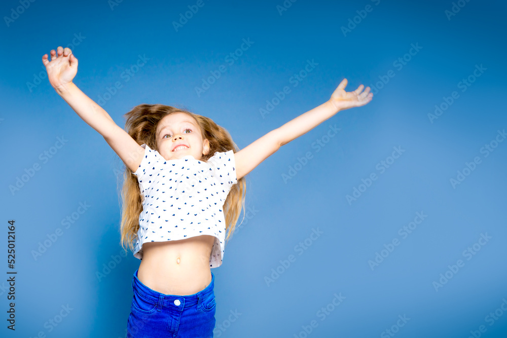 Cheerful little girl jumping lifts hands up. On a blue background in the studio Cheerful little girl jumping lifts hands up. On a blue background in the studio