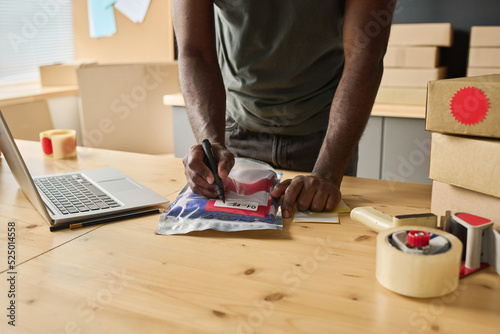 Close-up of African man making notes on packet with order at table while distributing parcels to addresses