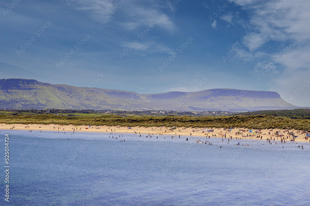 Busy Mullaghmore beach on a sunny day. Tourist swimming in the ocean and sunbathing on a sand. Benbulben mountain in the background. County Sligo, Ireland. Popular holiday area with stunning nature.