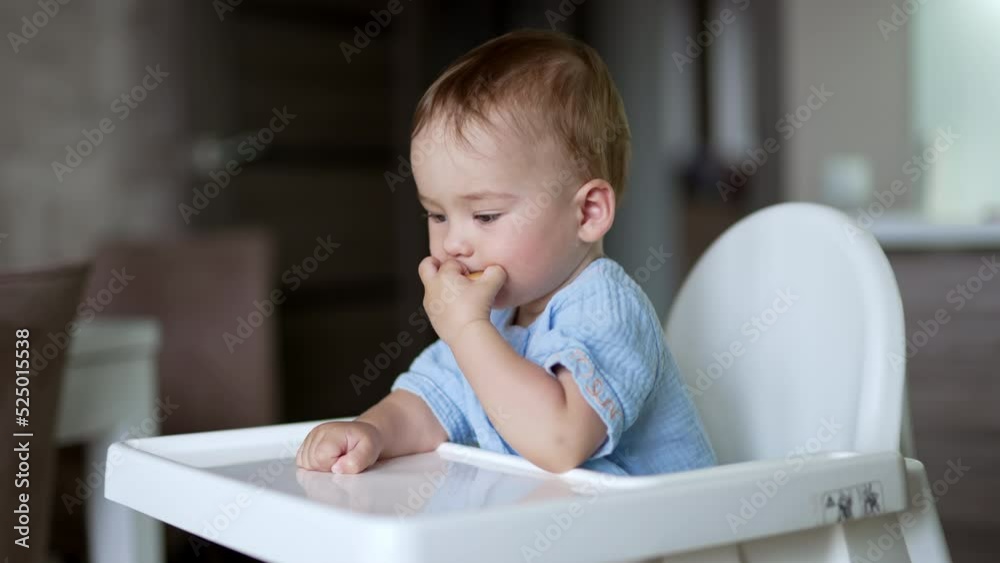 Little cute baby boy sitting at feeding table. Lovely child putting the bagel into mouth, throwing on the table and taking again. Side view. Little cute baby boy sitting at feeding table. Lovely child putting the bagel into mouth, throwing on the table and taking again. Side view.