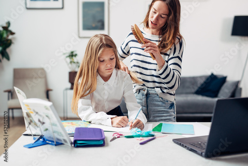 Mother brushes a girls hair while doing her homework.