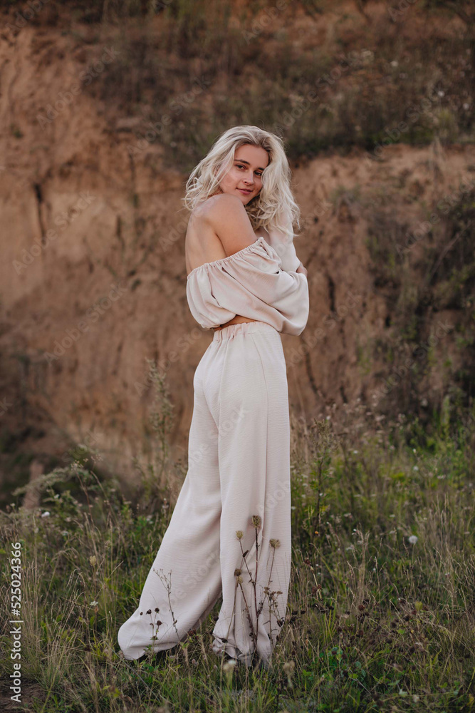A stylish blonde girl with tanned skin and curly hair stands in a beige suit with a white flower on an overgrown quarry. Fashion, travel and lifestyle concept.