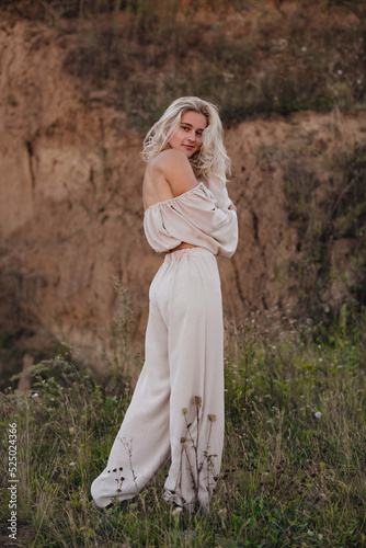 A stylish blonde girl with tanned skin and curly hair stands in a beige suit with a white flower on an overgrown quarry. Fashion, travel and lifestyle concept.