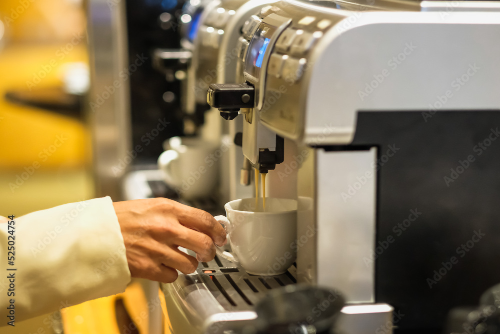 A hand using a coffee maker machine to brew an espresso coffee. Coffee being poured into white mug. A modern home office coffee machine appliance, selective focus in the banquet room A hand using a coffee maker machine to brew an espresso coffee. Coffee being poured into white mug. A modern home office coffee machine appliance, selective focus in the banquet room