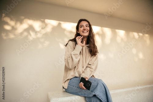 Cute young caucasian woman smiling looking away, holding smartphone sitting on white background. Brunette wears casual hoodie, jeans. Relaxation concept