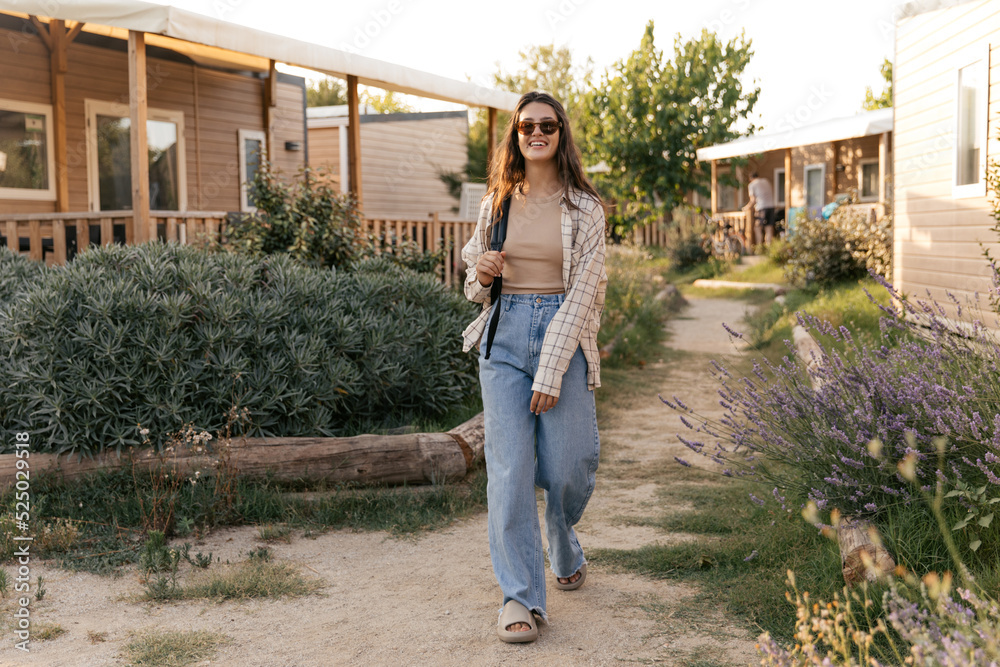 In full growth, nice young caucasian girl walks among cottages on vacation in spring. Brunette wears sunglasses, tank top, jeans, slippers and backpack. People sincere emotions lifestyle concept.