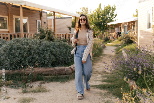 In full growth, nice young caucasian girl walks among cottages on vacation in spring. Brunette wears sunglasses, tank top, jeans, slippers and backpack. People sincere emotions lifestyle concept.
