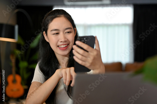 Attractive Asian female relaxes in comfortable living room using her smartphone