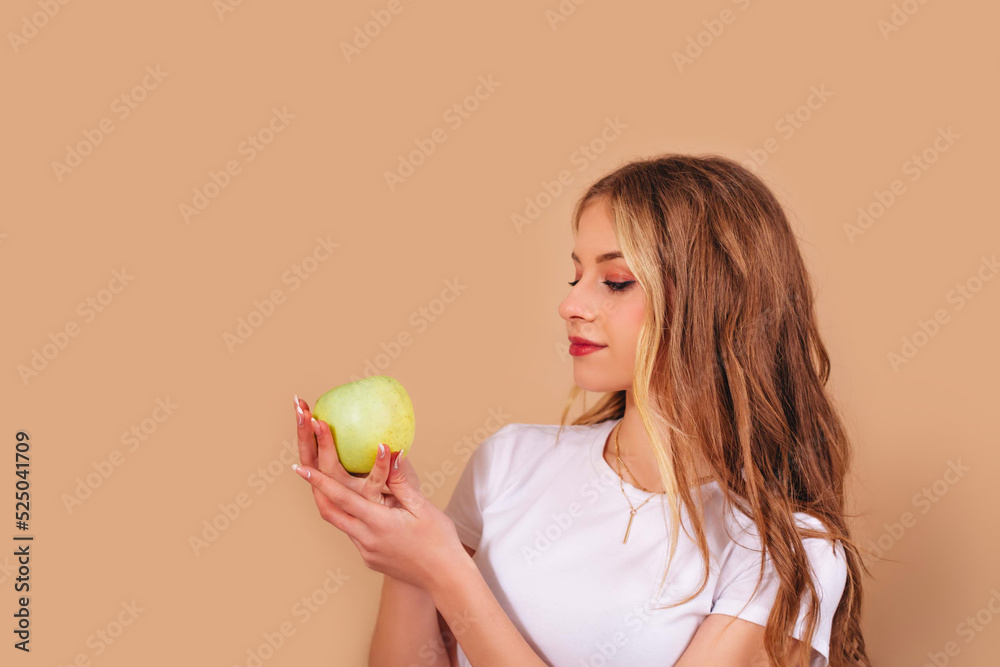 Teenage girl with long curly hair and in a white t-shirt holds a green apple in her hands isolated on a beige background. Copy space. Teenage girl with long curly hair and in a white t-shirt holds a green apple in her hands isolated on a beige background. Copy space.