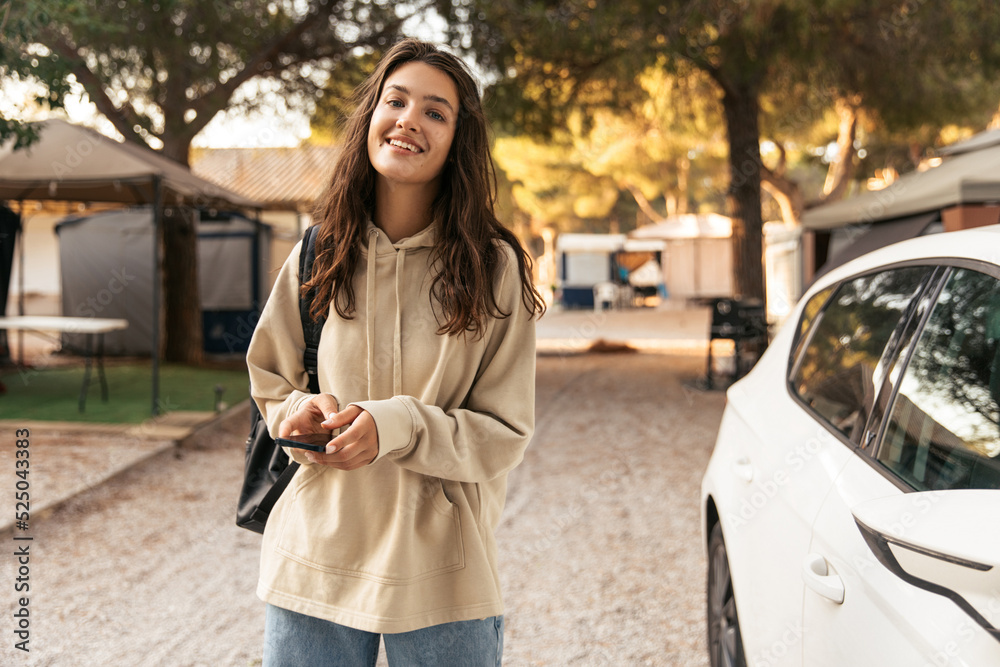 Fototapeta premium Nice young caucasian woman smiling looking at camera standing outdoors. Brunette wears hoodie, jeans and backpack. Leisure lifestyle and beauty concept.