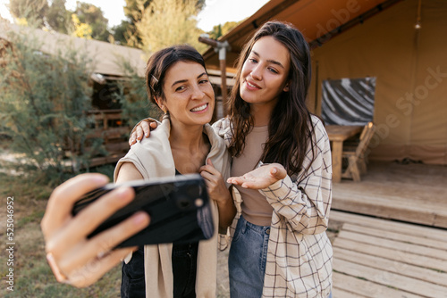 Attractive young caucasian ladies take pictures of themselves with help of phone spending vacations on street. Brunettes wear casual clothes in light colors. Lifestyle concept