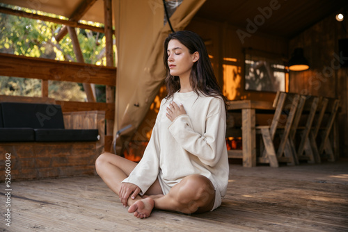 Focused young caucasian girl closing her eyes meditates in morning sitting on floor in campsite. Brunette wears homemade sweatshirt and shorts. Concept of harmony and tranquility