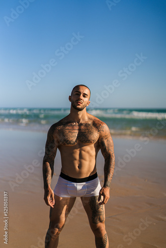 Chico joven tatuado y musculoso en ba  ador y ropa interior en la playa
