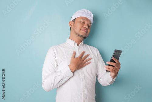 Happy mindful thankful young Balinese man holding phone and hand on chest smiling isolated on blue background feeling no stress, gratitude, mental health balance, peace of mind concept.
