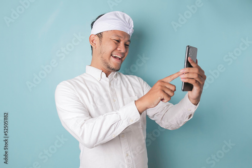 A portrait of a happy Balinese man is smiling and holding his smartphone wearing udeng or traditional headband and white shirt isolated by a blue background
