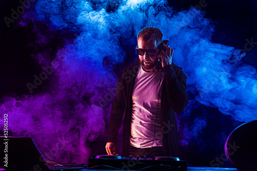 Young stylish man in glasses posing behind mixing console on colored smoke studio background