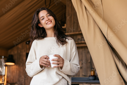 Pretty young caucasian woman in good mood spends her free time alone indoors. Brunette is looking at camera, holding cup of tea. Recreation concept #525052748