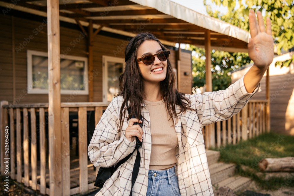 Cheerful young caucasian girl waving her hand looking away relaxing outdoors. Brunette wears sunglasses, tank top, shirt and backpack. Leisure lifestyle and beauty concept.