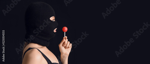 Obraz na płótnie Young woman in balaclava and with lollipop on black background with space for te