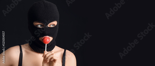 Fotografia Young woman in balaclava and with lollipop on black background with space for te