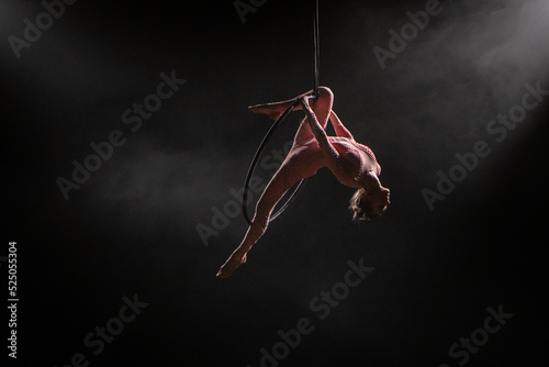 Aerial acrobat in the air ring. Young woman performs the acrobatic elements in the air hoop. Aerialist in on black background dark studio with backlight and smoke. For sports, acrobatic, circus school