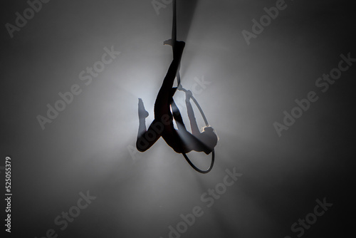 Silhouette of young woman gymnast performs acrobatic stunts on an air hoop under the dome of a circus. Exciting acrobatic show in a dark smoky studio with backlight. photo