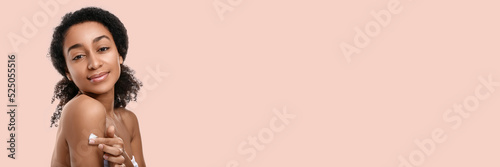 Young African-American woman applying body cream against beige background with space for text