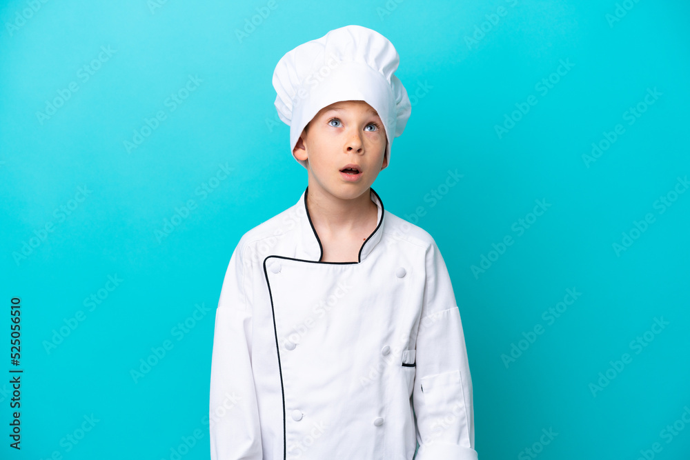 Little chef boy isolated on blue background looking up and with surprised expression Little chef boy isolated on blue background looking up and with surprised expression