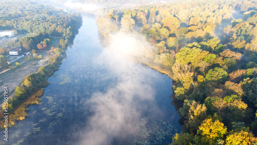 Aerial drone view flying over the river with calm reflective water surface and white fog mist and forest trees on the banks of the river on a sunny early autumn morning. Beautiful natural background.
