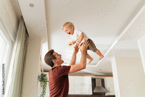 Young dad throwing excited laughing baby boy up in air and catching, amusing kid and having fun together after returning home from work while mother cooking dinner. Active play at home photo