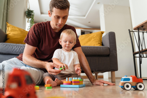 Father assisting his baby boy playing sorter sitting on floor in living-room with sofa on background, surrounded with car toys, waiting mom to come home from work. Education toys, child development