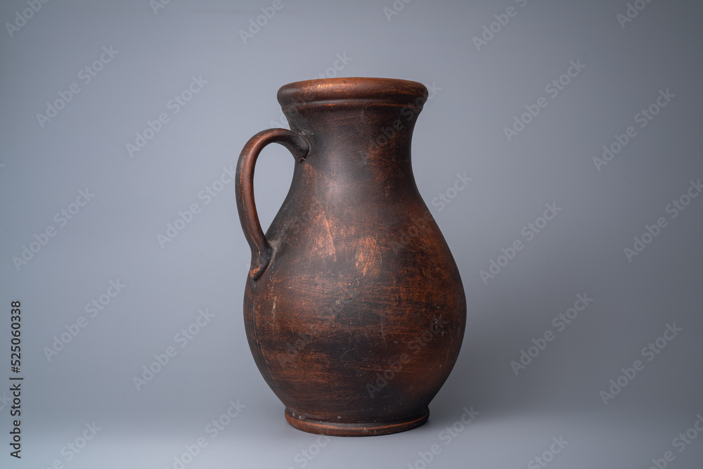 Dark brown jug with a handle, handmade jar from clay on a gray background in the studio. Earthenware for drink and food. Kitchen utensils. Old rustic cookware made of earthen. Retro craft vessel.