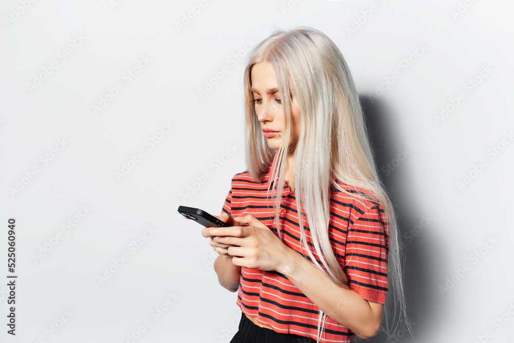 Studio portrait of young pretty girl with blonde hair, looking in smartphone, wearing red striped shirt on white background. Studio portrait of young pretty girl with blonde hair, looking in smartphone, wearing red striped shirt on white background.