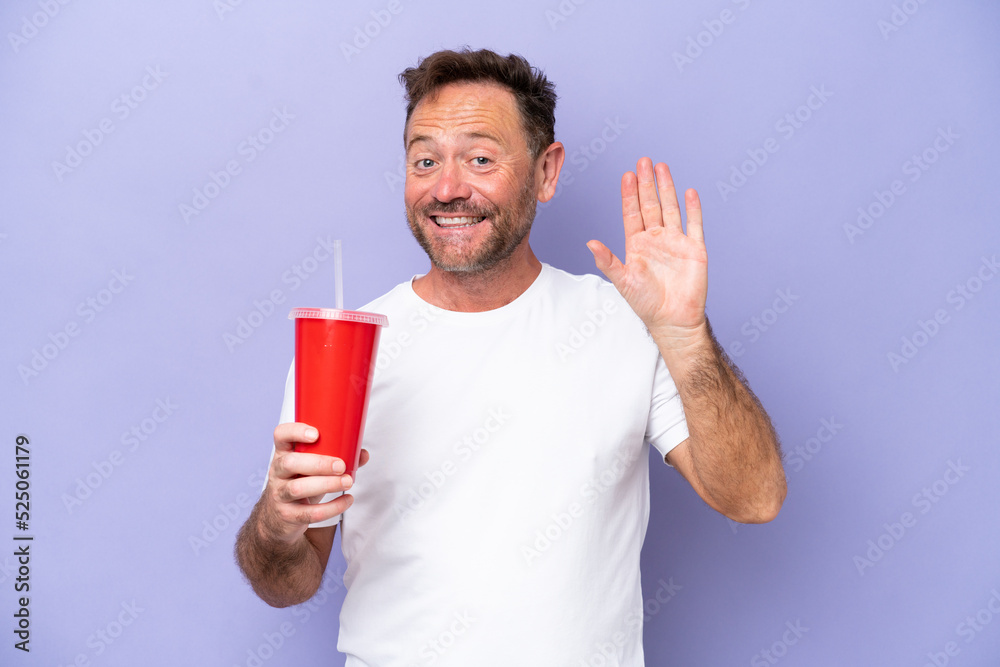 Middle age caucasian man holding soda isolated on purple background saluting with hand with happy expression