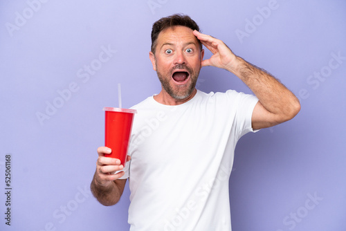 Middle age caucasian man holding soda isolated on purple background with surprise expression © luismolinero