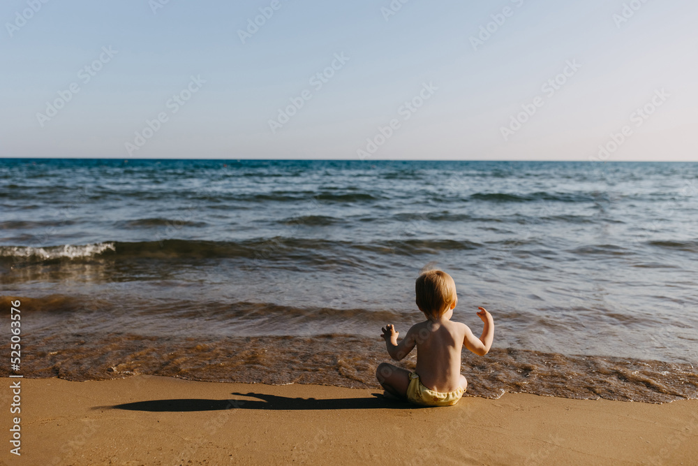 Little child sitting alone on a seashore, watching waves