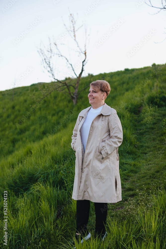An aged woman with a short haircut and in a beige raincoat walks on a hill in autumn.