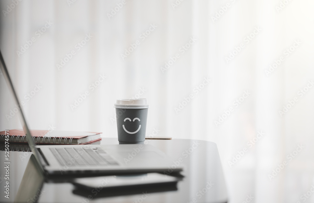 smiling face on paper cup of hot coffee with laptop on desk besides window