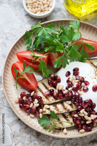 Grilled aubergine slices with pomegranate, pine nuts, white yogurt, tomatoes and parsley on a beige plate, close-up, vertical shot