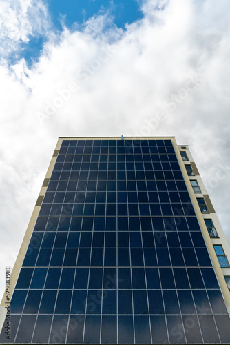 A modern energy-efficient building against the background of clouds. Multi-storey residential building with solar panels on the wall. Renewable energy sources in the city