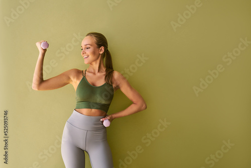 Attractive sporty woman wearing green top and gray leggings standing near green background holding pink dumbbells and smiling.