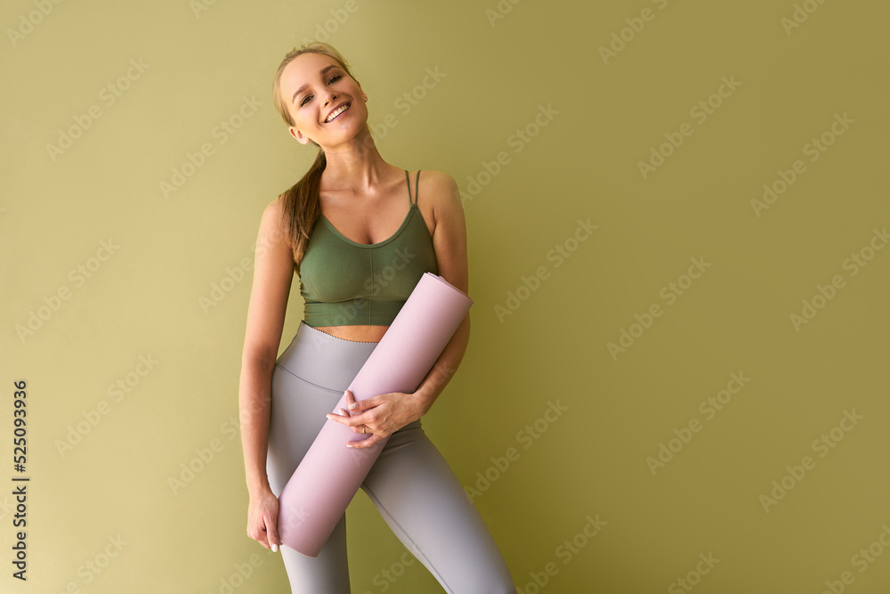 Attractive sporty woman wearing a green top and gray leggings stands near a  green background holding a pink sports mat and smiling. Stock Photo