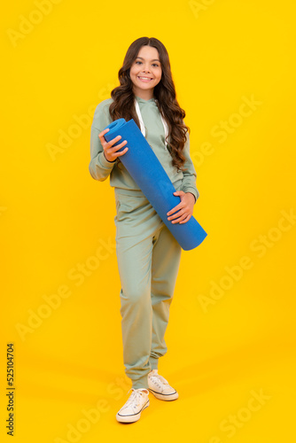 Full length of a fitness teen girl in sportswear hold yoga mat posing over yellow background. Fitness model child wearing sport clothes. Girl in the sport concept.