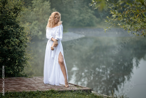 a red-haired girl stands in a white dress on the shore of a lake at dawn