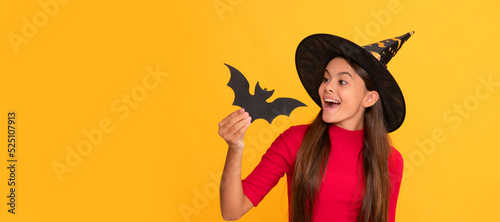 positive teen girl wearing witch hat holding bat on yellow background, childhood. Halloween kid girl portrait, horizontal poster. Banner header with copy space.