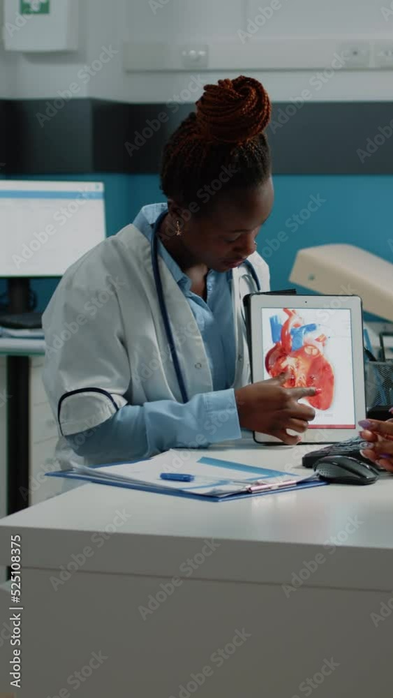 Vertical video: Doctor showing tablet with heart figure for cardiology checkup to patient at consultation. Young parent looking at device with organ for cardiovascular and blood flow healthcare. Vertical video: Doctor showing tablet with heart figure for cardiology checkup to patient at consultation. Young parent looking at device with organ for cardiovascular and blood flow healthcare.